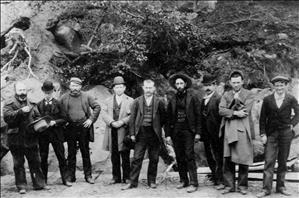 Nine men stand at the base of a sandy bluff. Some of them are wearing floppy hats. 
